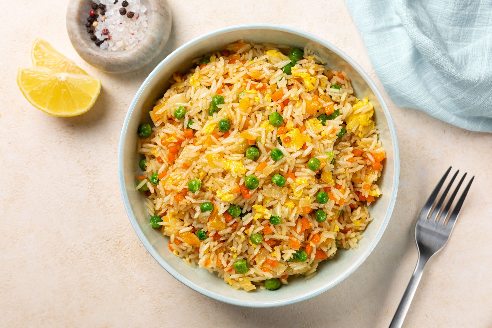 Top view of a bowl with fried rice with egg and vegetables, onion, bell pepper, carrot, green peas. Vegetarian food served in a light blue plate on a beige table.