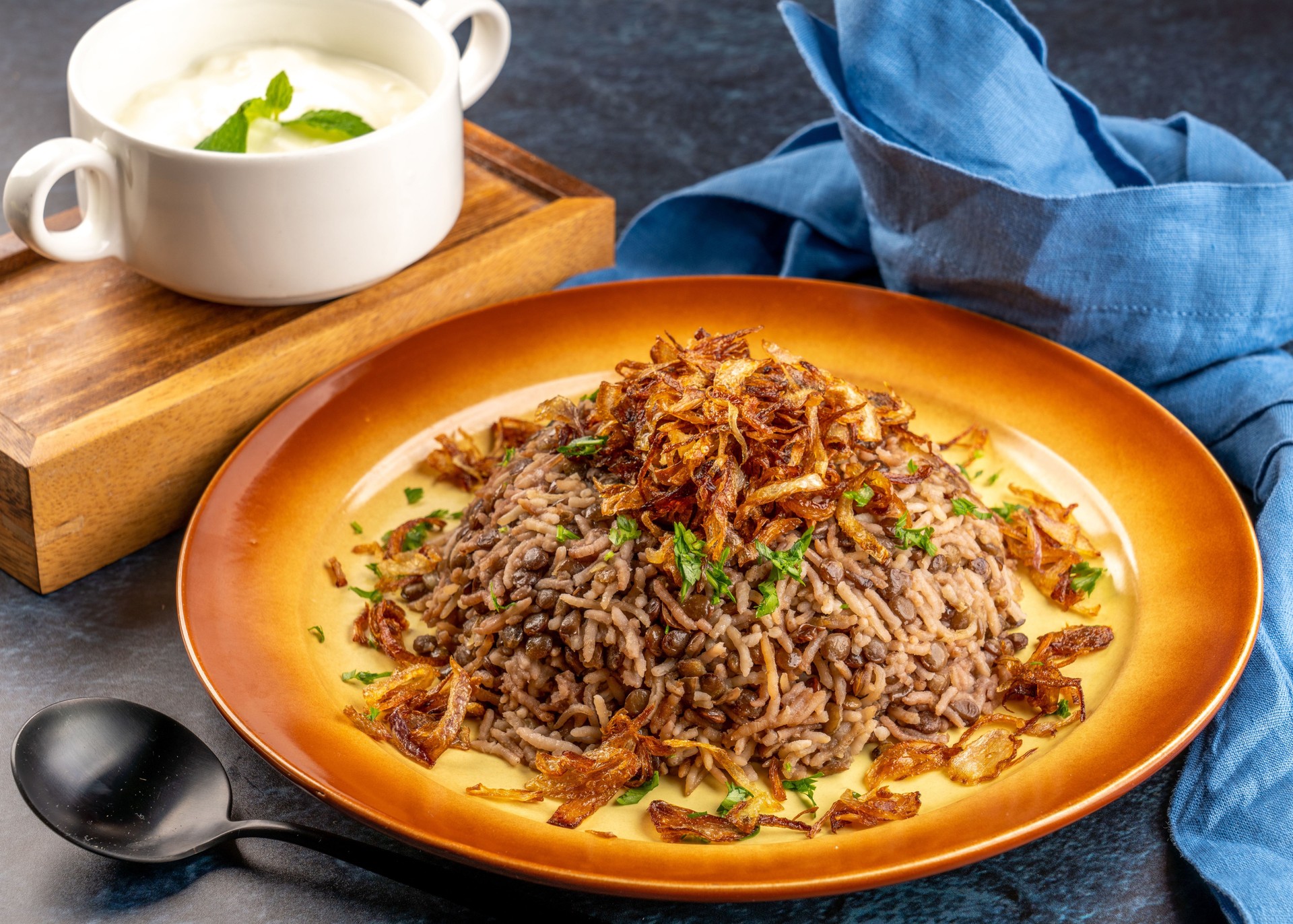 Closeup shot of brown rice with fried onions and a bowl of yogurt on a wooden tray
